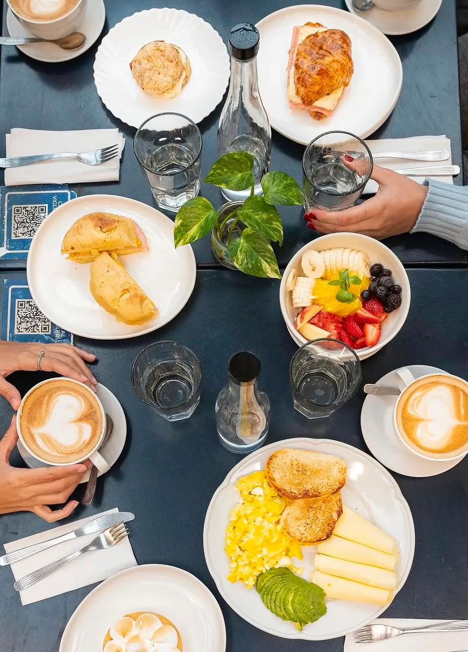 Mesa de desayuno vista desde arriba: cafés con leche, huevos revueltos, tostadas, medialunas y bowl de frutas